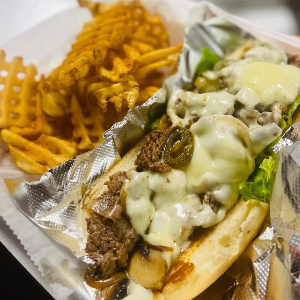 Cheesesteak with melted cheese, jalapeños, and lettuce, served with waffle fries in a foil-lined basket.
