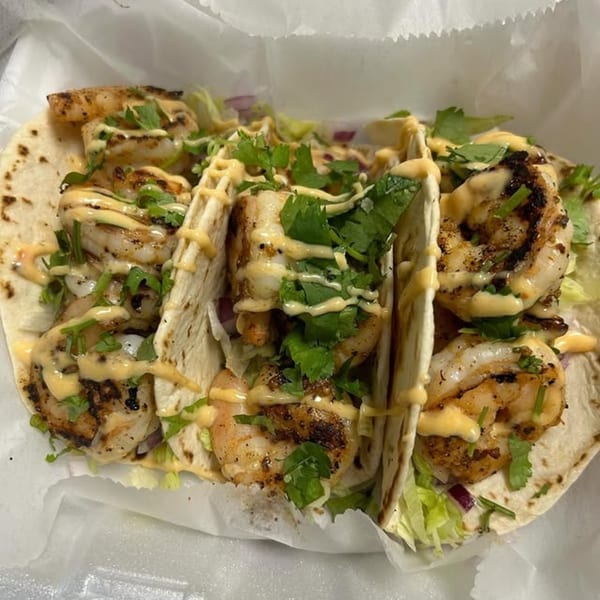 Three grilled shrimp tacos with cilantro, lettuce, and a creamy sauce in a white paper container.