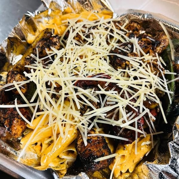 Soul food dish with grilled chicken, creamy pasta, and shredded cheese in a foil container.